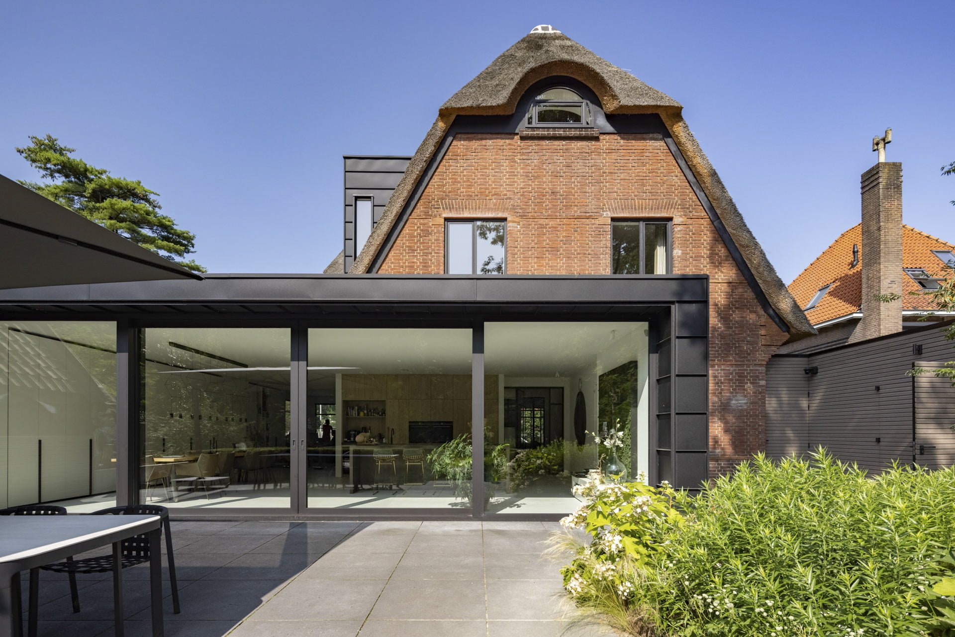 Vierdelige zwarte Aluprof aluminium schuifpui in moderne aanbouw tegen klassieke woning met rieten dak – Toelevering Online