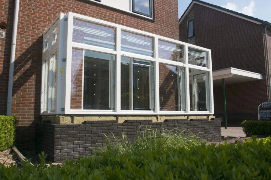 Woning met serre-uitbouw voorzien van witte kozijnen en grote glaspartijen, geplaatst op verhoogde bakstenen plint – Toelevering Online